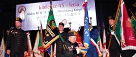 Na scenie stoją ludzie na baczność obok sztandarów. Jeden Pan klęka i trzyma w ręce kawałek sztandaru powiatu polkowickiego.