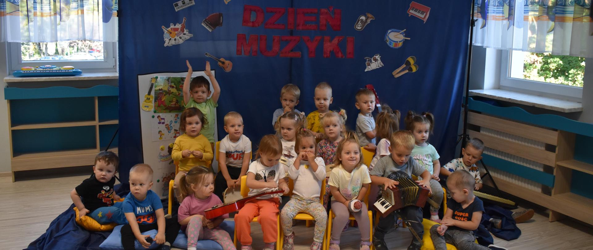 Na zdjęciu widać dzieci z grupy Delfinki pozujące do grupowego zdjęcia. Za dziećmi granatowe tło z napisem Dzień Muzyki.