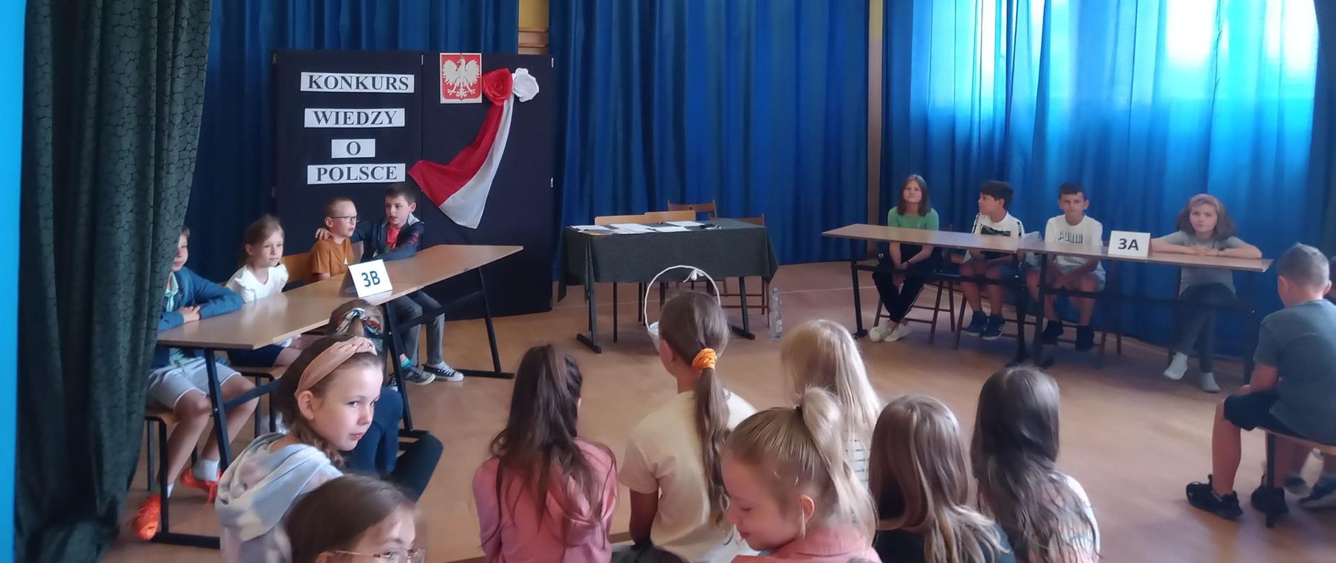 Dzieci w dwóch grupach siedzą przy stolikach. Za nimi ściana z granatowych zasłon