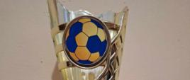 Złoty puchar z zamieszczoną na nim złoto-niebieską piłką za pierwsze miejsce w Gminnym Turnieju Piłki Ręcznej w kategorii chłopcy