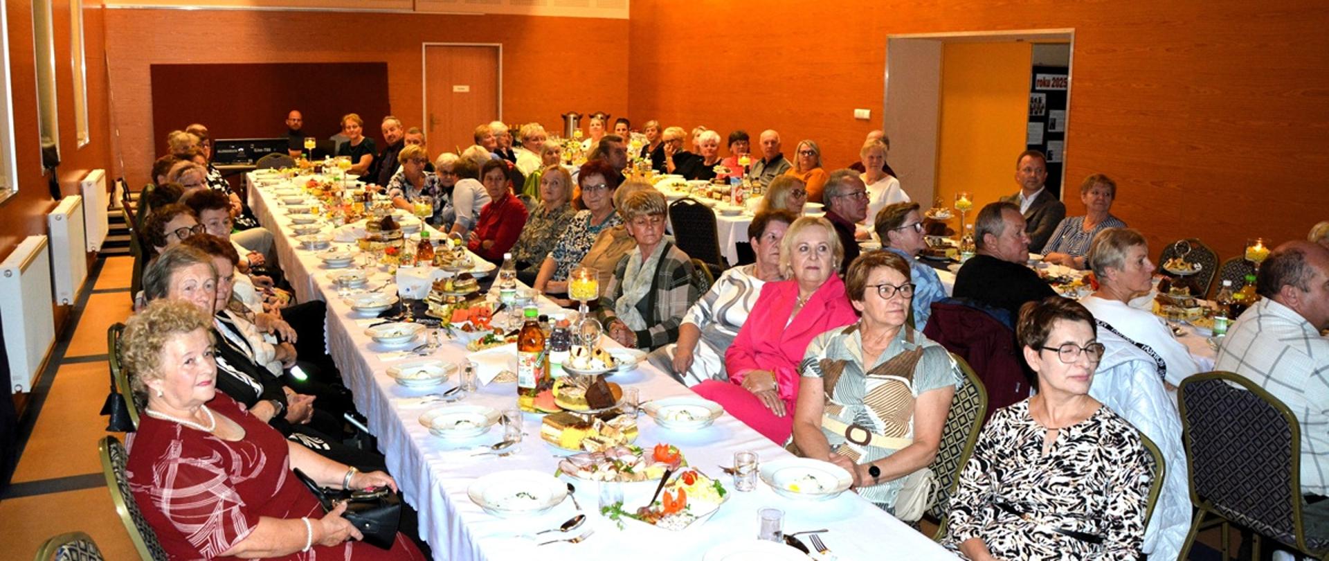 kobiety i mężczyźni odświętnie ubrani siedzą przy suto zastawionych stołach 