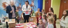 Pani Wójt Gminy Łabowa Marta Słaby oraz Pan Dawid Madziar, Prezes Fundacji Socius, wręczają uczniom klas 1-3 Szkoły Filialnej w Roztoce Wielkiej nagrody za udział w konkursie plastycznym w ramach projektu "W sercu Ojczyzny", na stoliku leżą prace plastyczne uczniów, w tle dekoracja ze świętym Mikołajem, po prawej stronie uczniowie odbierający nagrody