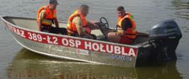 trzej mężczyźni w kamizelkach ratunkowych siedzących w zwodowanej łodzi, z napisem "Straż 389-Ł2 OSP Hołubla"