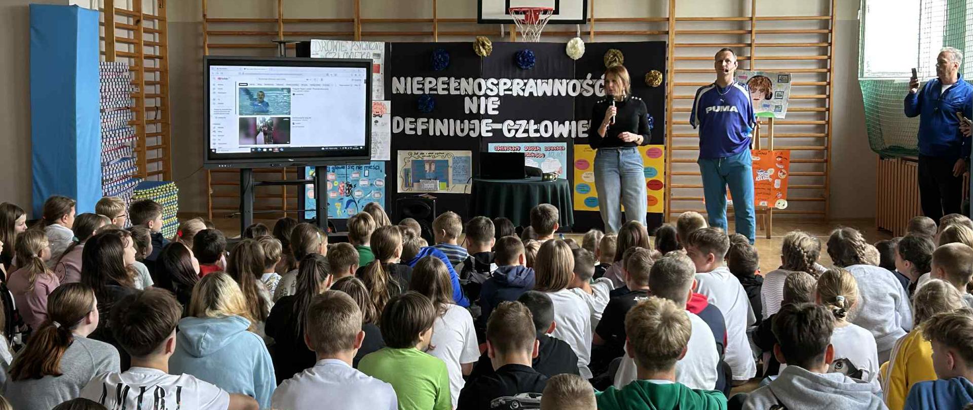 Dzieci siedzące w dużej liczbie. Przed nimi stoi kobieta z mikrofonem w prawym ręku i mężczyzna bez rąk. Za nimi biały napis Niepełnosprawność nie definiuje człowieka