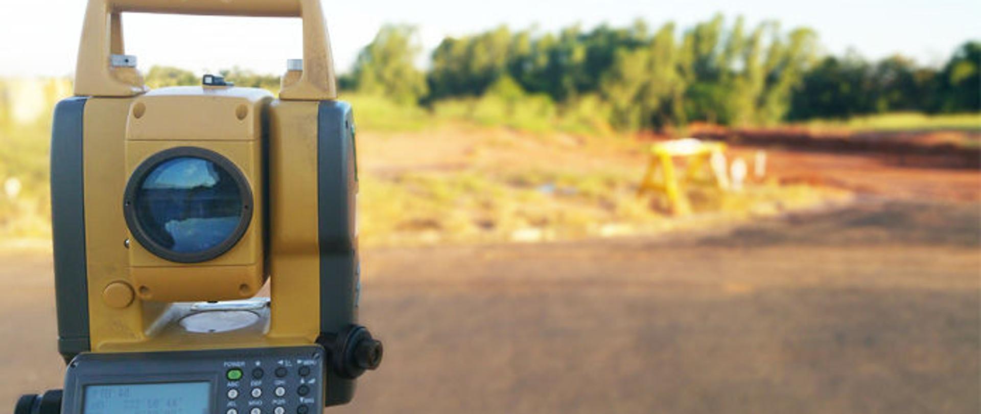 Teodolit elektroniczny stoi na tle działek i lasu