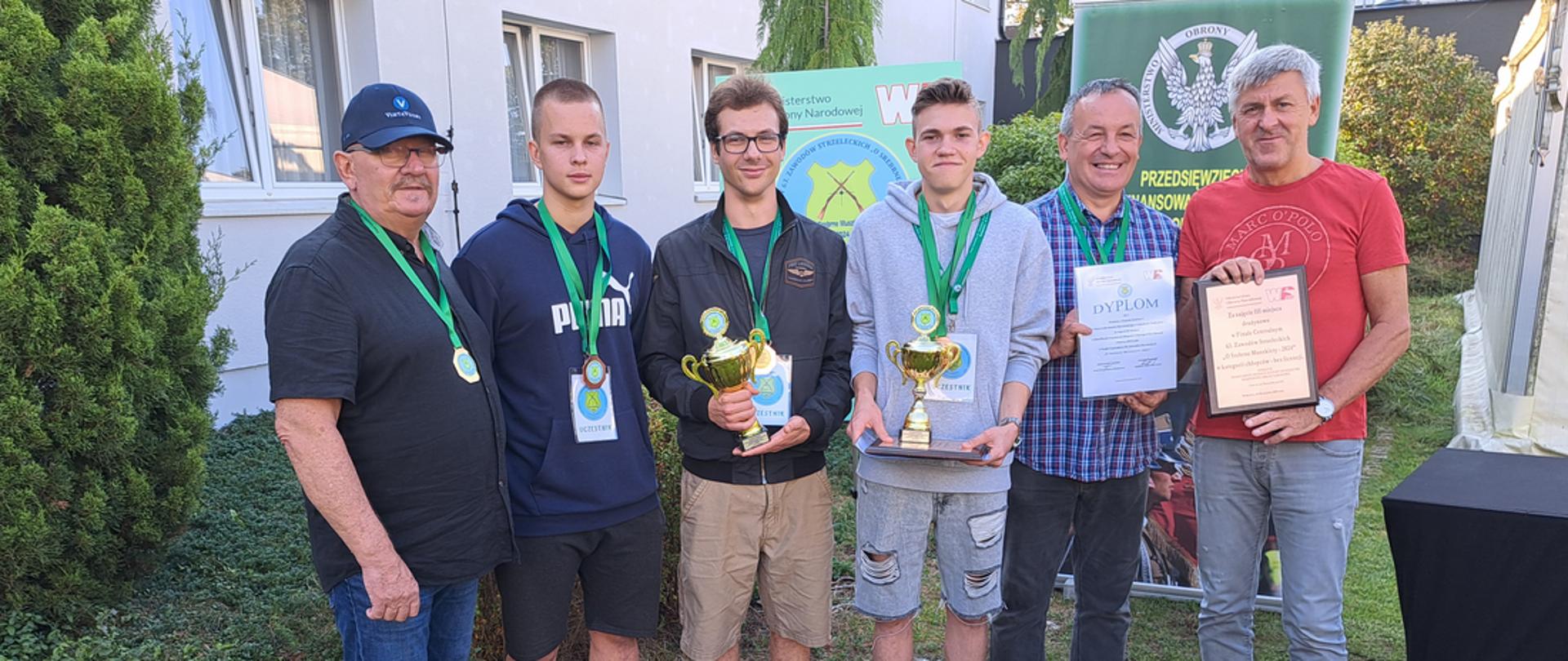 Na zdjęciu widzimy Drużynę Zespołu Szkół Nr 1 wraz z organizatorami, która prezentuje zdobyte trofea (dyplomy i puchary) w tle budynek oraz przylegająca do niego zieleń.