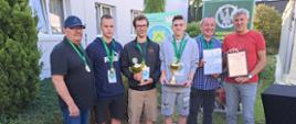 Na zdjęciu widzimy Drużynę Zespołu Szkół Nr 1 wraz z organizatorami, która prezentuje zdobyte trofea (dyplomy i puchary) w tle budynek oraz przylegająca do niego zieleń.