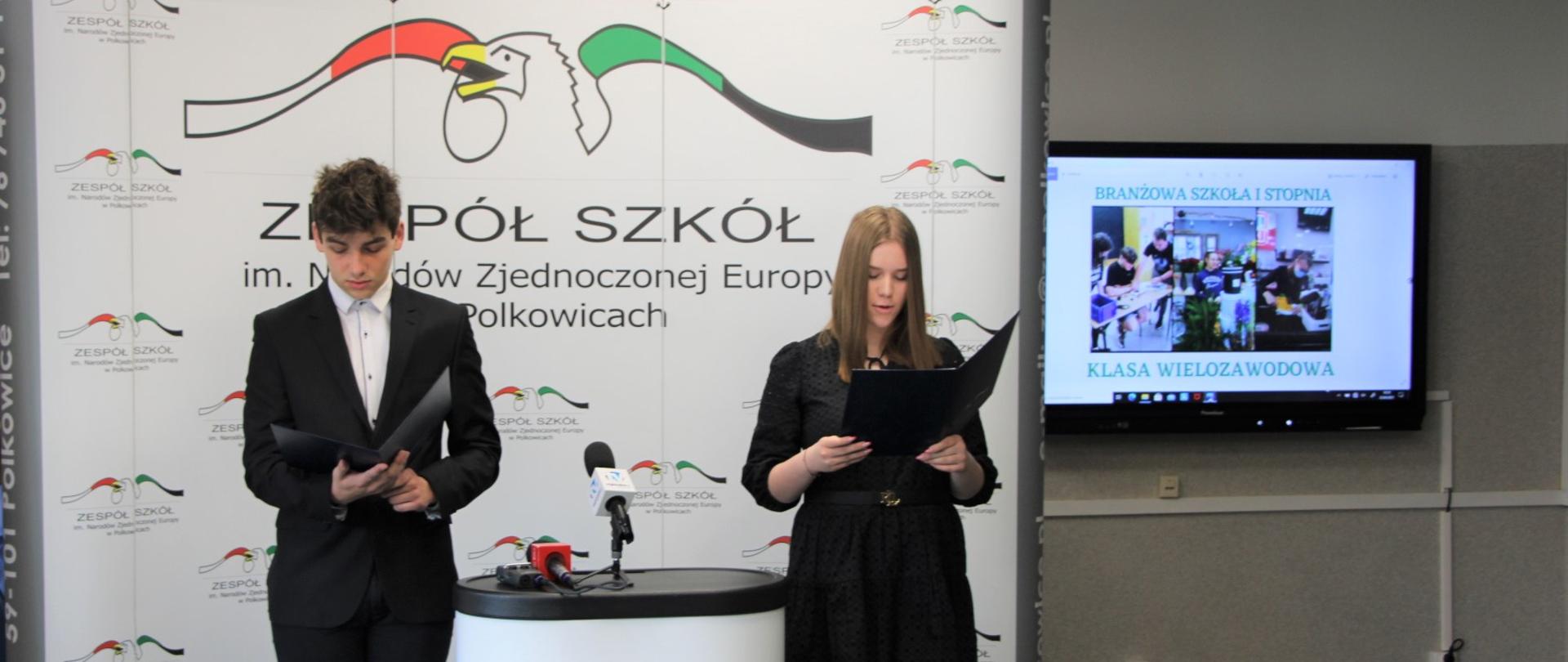 Na tle ścianki ekspozycyjnej z logo Zespołu Szkół im. Narodów Zjednoczonej Europy stoją elegancko ubrani uczniowie. Pomiędzy nimi stoi stolik z mikrofonami.