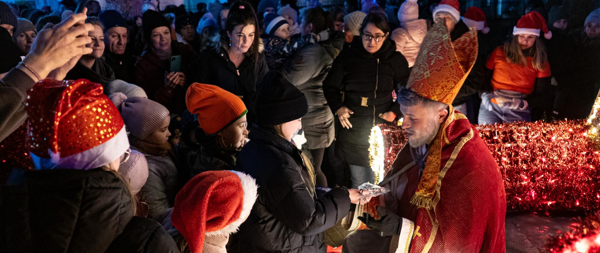 Wizyta św. Mikołaja na Rynku w Kalwarii Zebrzydowskiej. Obok Mikołaja znajduje się duża ilość dzieci i starszych mieszkańców, którzy oczekują na rozdanie przez niego prezentów.