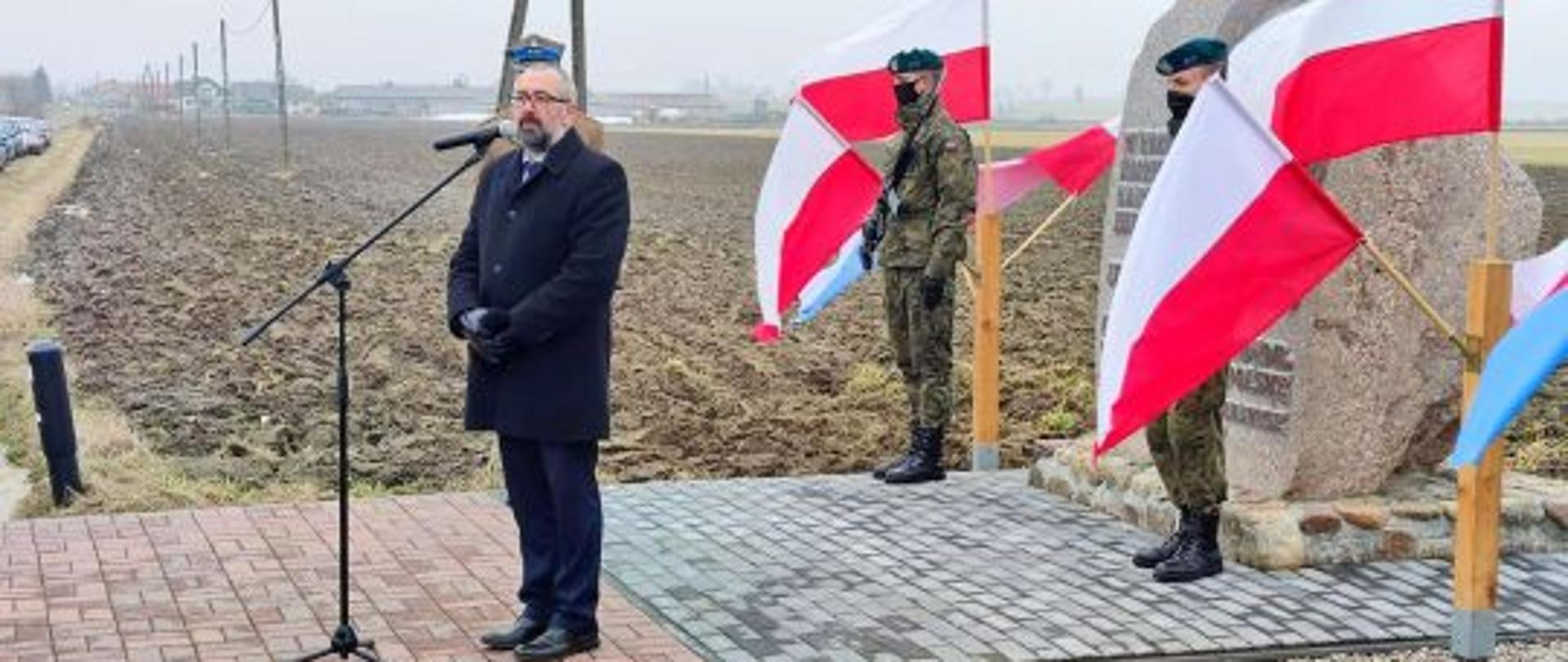 Starosta Krzysztof Bieńkowski przemawia do zgromadzonych w Dniu Pamięci Żołnierzy Wyklętych w Gostkowie. Za Starostą Przasnyskim pomnik poległych Żołnierzy Wyklętych ze składu patrolu Edwarda Dobrzyńskiego „Orzyca”, warta honorowa żołnierzy 2. Ośrodka Radioelektronicznego w Przasnyszu i łopoczące biało czerwone flagi.