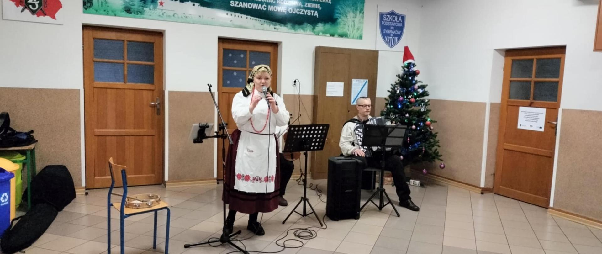 Kobieta ubrana w strój ludowy śpiewa przez mikrofon. Za nią siedzi mężczyzna trzymający na kolanach akordeon.