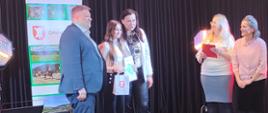 Nagrodzona uczennica trzyma w ręku nagrodę oraz dyplom, pozuje do zdjęcia z panią wójt i wicewójtem, obok stoją 2 panie organizatorki konkursu