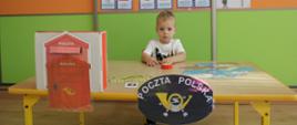 Na zdjęciu widać chłopca przyklejającego znaczek pocztowy do listu. Przed chłopcem znajduje się ilustracja z logo Poczty Polskiej oraz stoi skrzynka na listy. W tle znajdują się kolorowe obrazki przedstawiające listonosza, list, znaczek pocztowy, paczkę.