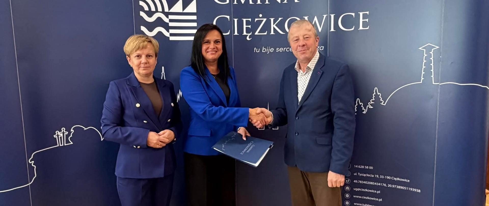 Trzy osoby siedzące na tle granatowej ściany z napisem gmina Ciężkowice