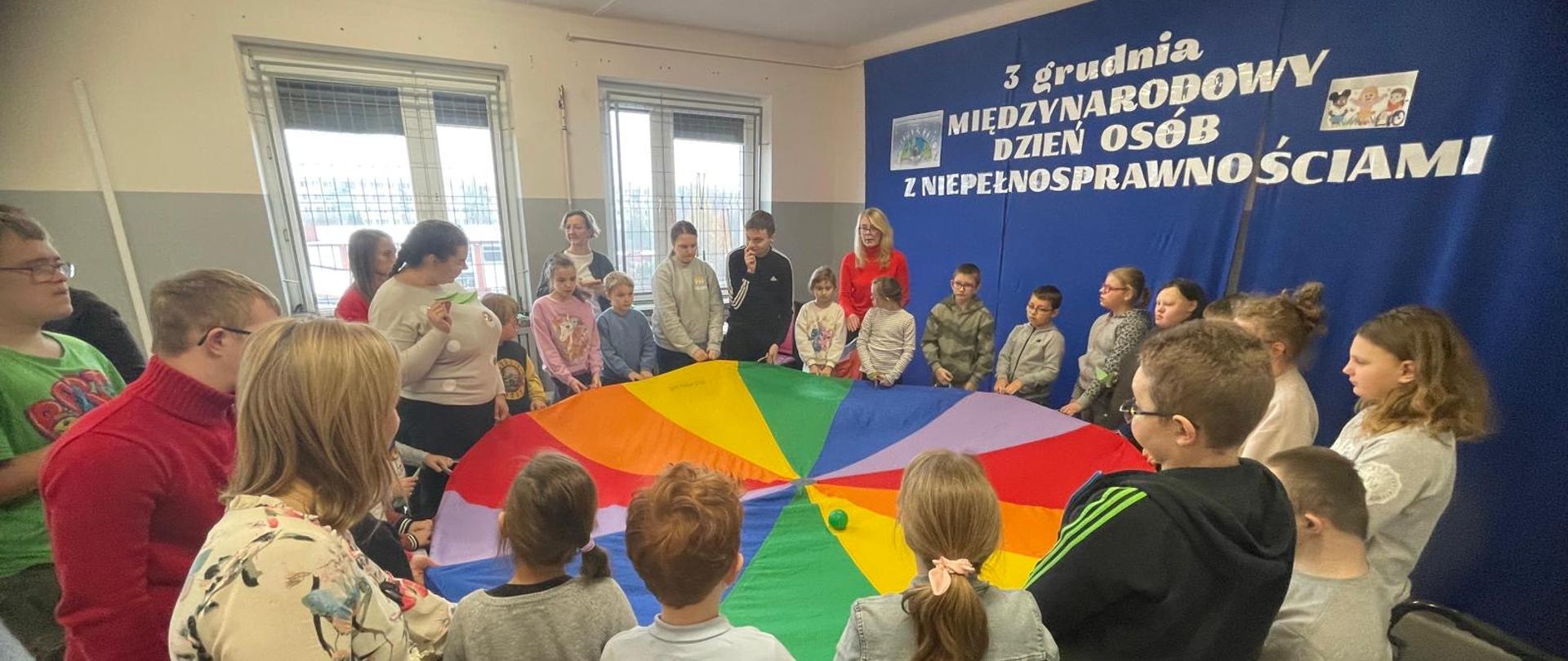 Na zdjęciu widzimy Panią z Biblioteki, która przeprowadza zabawę z chustą klanza. Dzieci podrzucają piłeczki.
