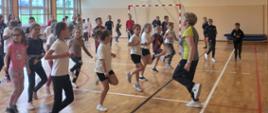 Uczniowie biegają w miejscu na sali gimnastycznej, przed nimi nauczycielka wychowania fizycznego pokazuje ćwiczenia 