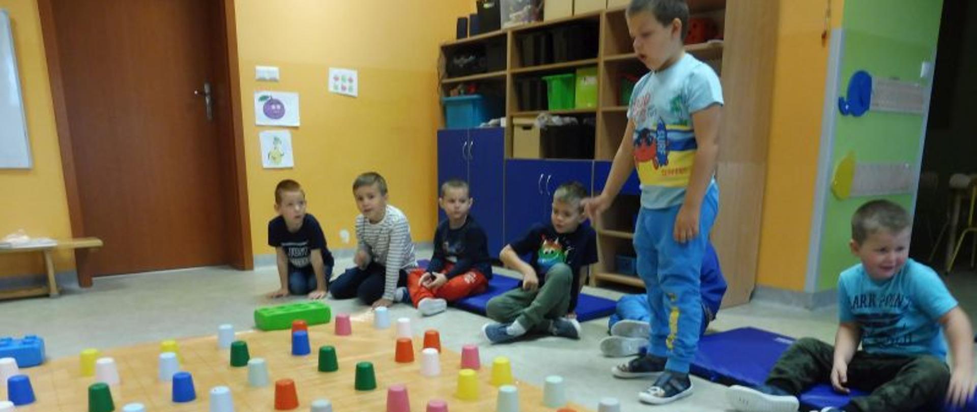 Sześć chłopców siedzi na materacach, jeden stoi przed matą na której są poukładane odwrócone do góry dnem kubeczki. 