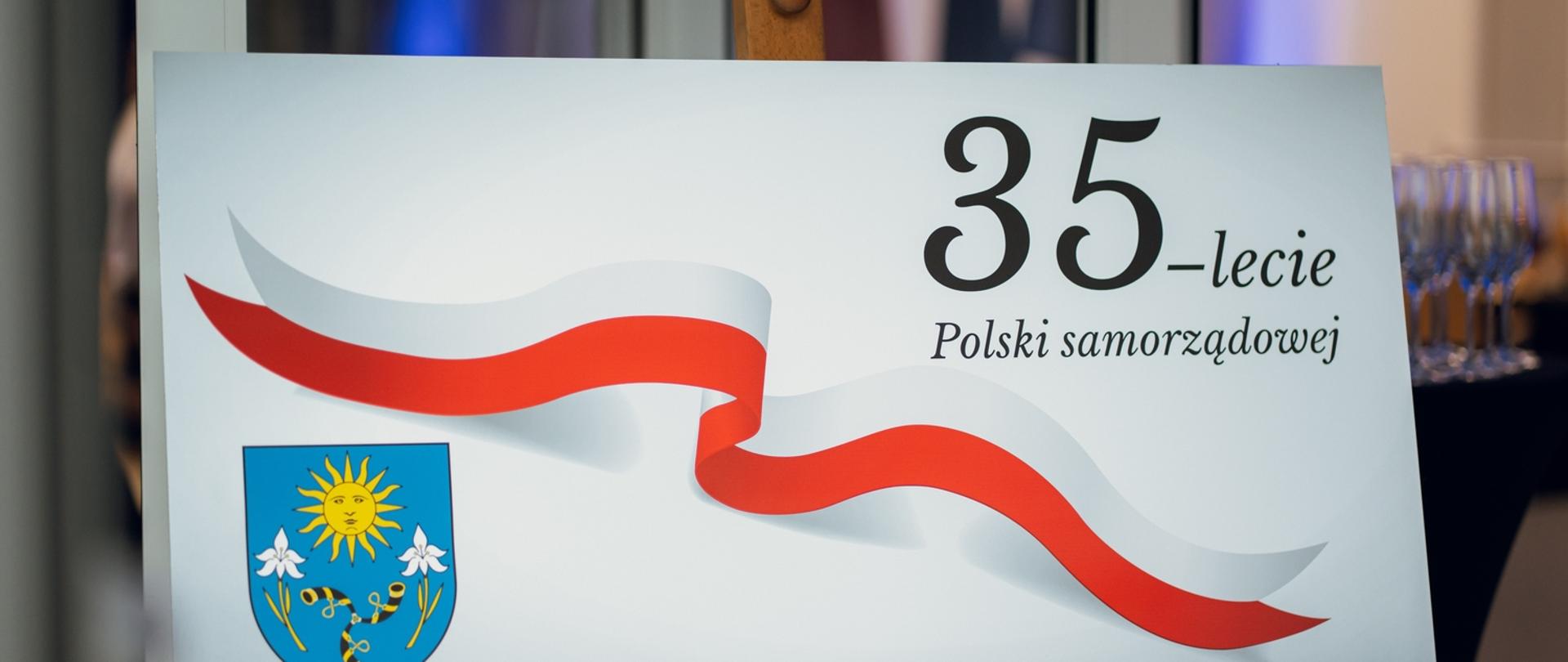 Plansza ustawiona na sztaludze, z grafiką biało-czerwonej wstęgi i napisem „35-lecie Polski samorządowej”. W lewym dolnym rogu widoczny herb z motywem słońca i kwiatów na niebieskim tle.