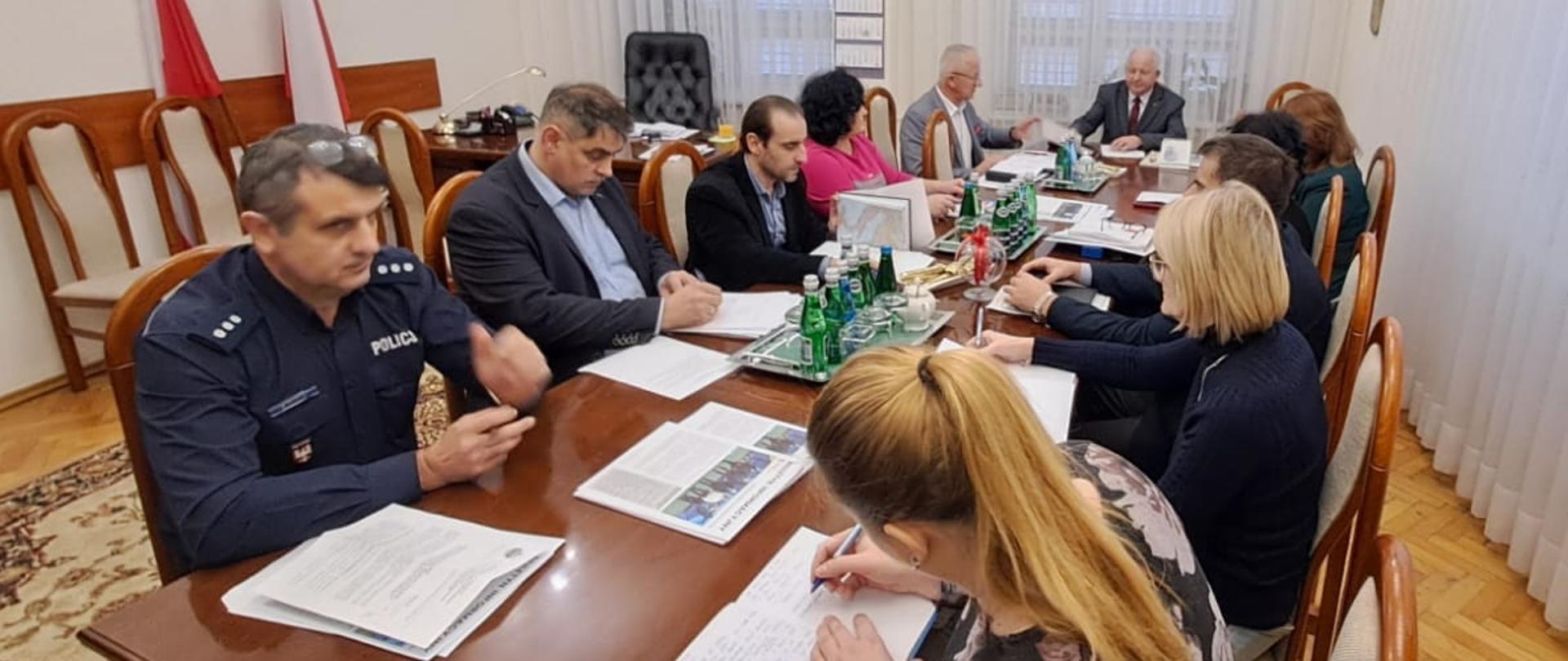 Przy stole konferencyjnym siedzi grupa jedenastu osób debatując. Przed nimi porozkładane materiały prasowe. 