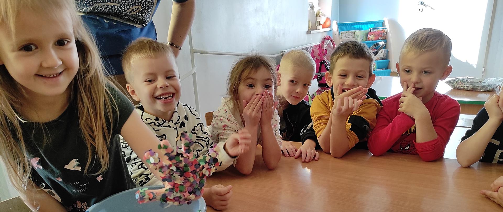 Zdjęcie przedstawia uśmiechnięte dzieci. Na stoliku znajdują się dwa pojemniki- jeden pusty, drugi z kolorowymi wydzierankami.