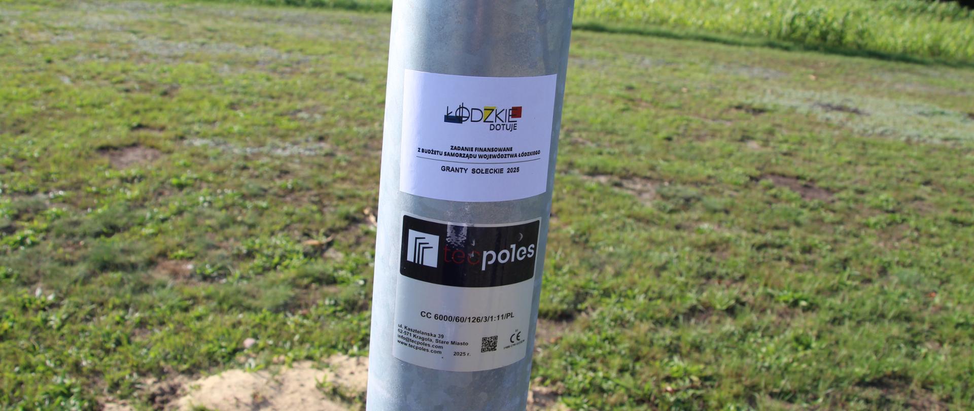 Bardzo bliskie ujęcie (detal) słupa latarni, na którym umieszczone są dwie naklejki informacyjne. Górna, mniejsza, biała naklejka zawiera logo „ŁÓDZKIE DOTUJE” oraz informację o "GRANTY SOŁECKIE 2025". Poniżej znajduje się czarna naklejka z logo producenta słupów "polsk poles" oraz kolejna, dolna, z kodami certyfikacji (np. CC 6000/60/126/1711MPL) i znakiem CE. Słup jest srebrny. W tle widać zieloną, wysoką trawę, pola uprawne (prawdopodobnie kukurydza) oraz ciemnozielony las na horyzoncie.
