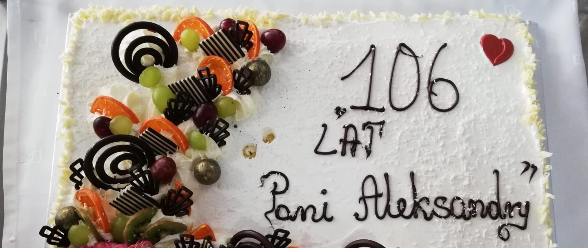 Tort z napisem: "106 lat Pani Aleksandry", na torcie znajdują się ozdoby z czekolady i owoców.