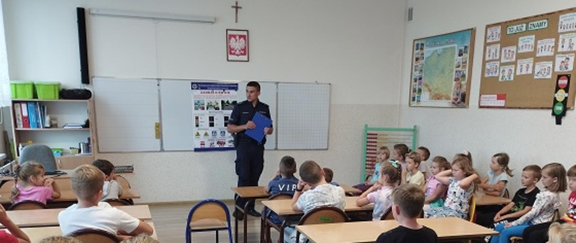 Dzieci siedzą w ławkach i słuchają prelekcji policjanta.