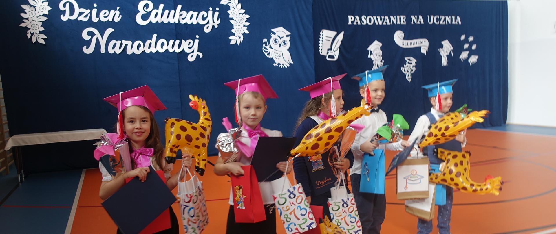 Uczniowie klasy I obdarowani prezentami stoją na tle dekoracji 