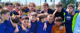 Zawodnicy z klas 4-6 w niebieskich strojach i z klas 7-8 w granatowo-zielonych strojach stoją obok siebie obejmując się ramionami, świętują zwycięstwo