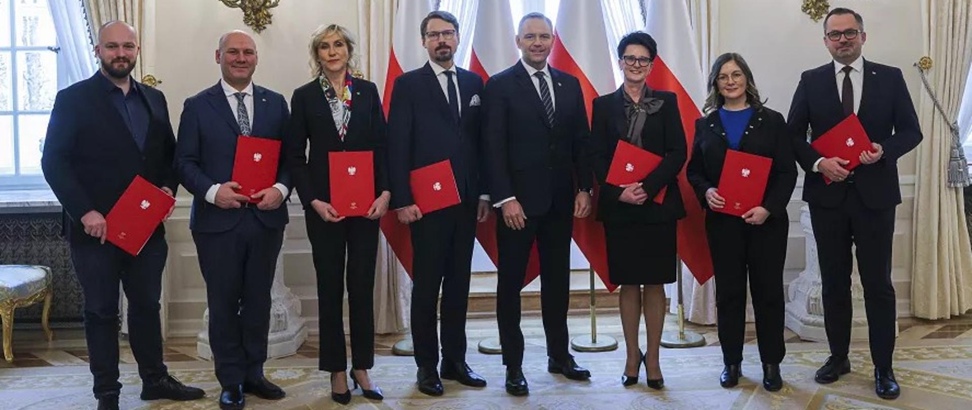 Grupa osób w białej sali pozuje do zdjęcia trzymając czerwone teczki.