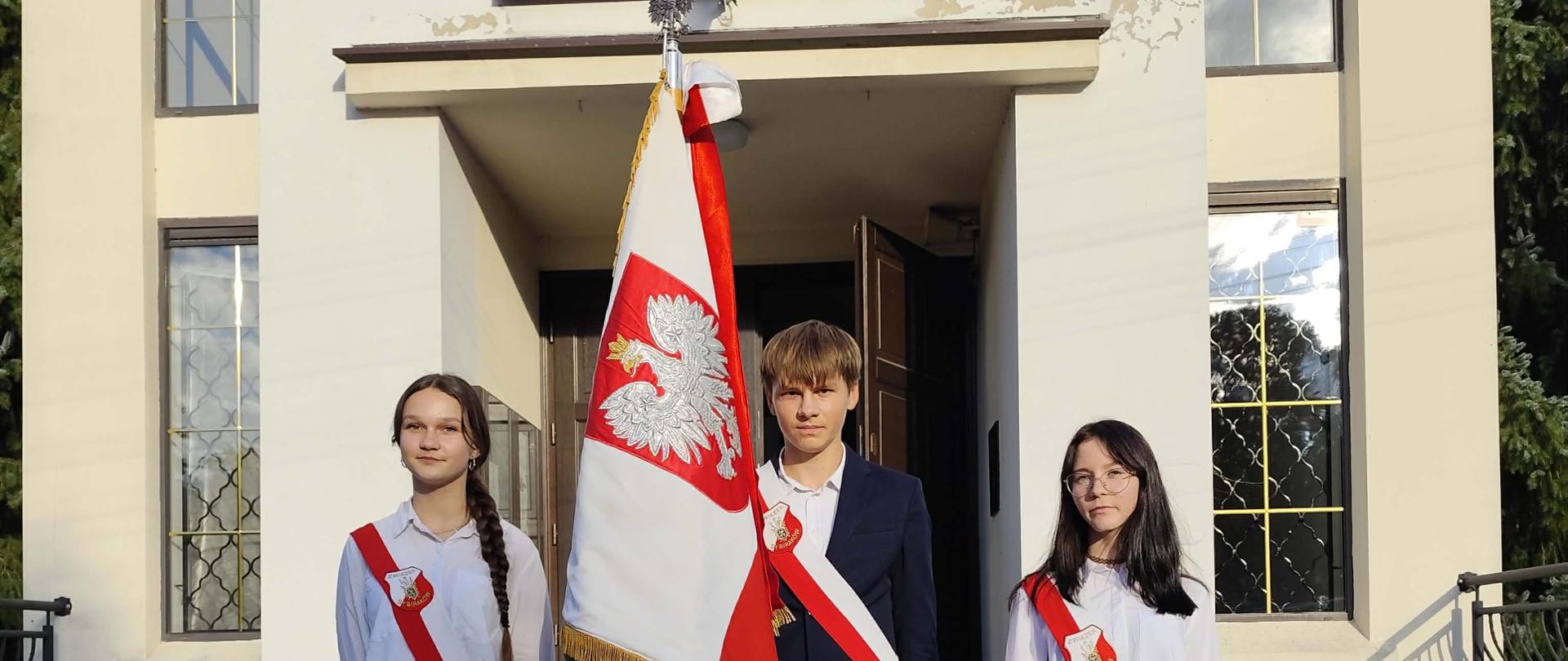 Zdjęcie przedstawia trzyosobowy szkolny poczet sztandarowy – dwie dziewczyny i chłopca – stojący na schodach przed wejściem do kościoła.