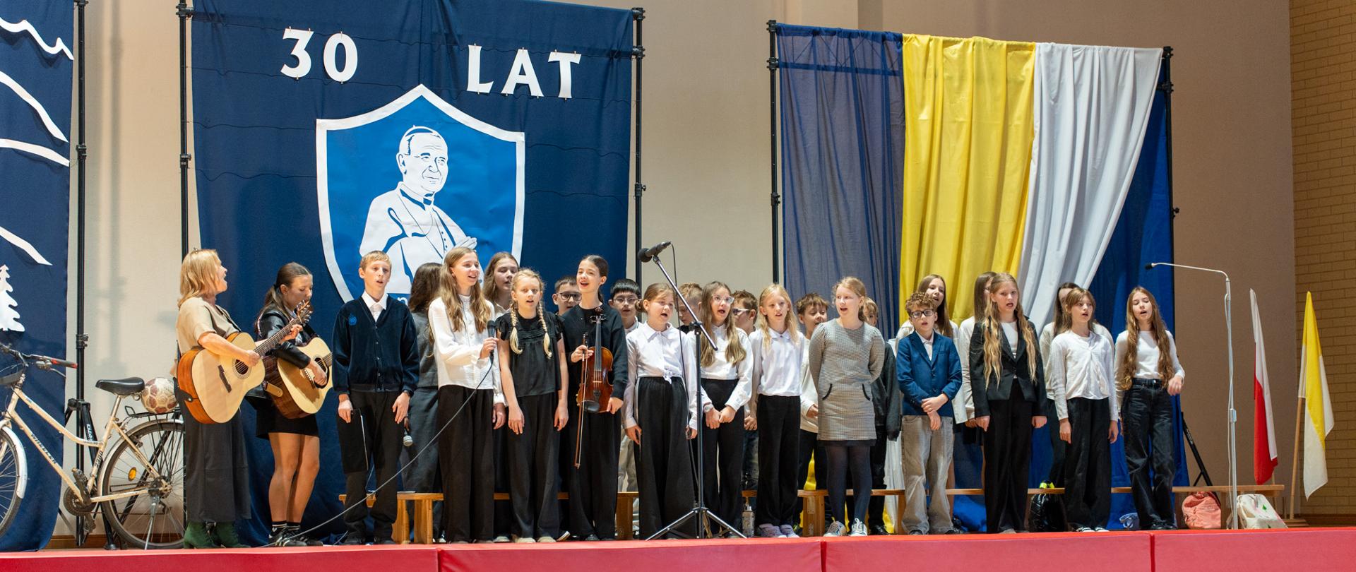 Zdjęcie przedstawia występ artystyczny uczniów Szkoły Podstawowej im. św. Jana Pawła II w Stanisławiu Dolnym podczas uroczystości z okazji 30-lecia nadania szkole imienia patrona, która odbyła się 22 października 2026 roku. Na scenie grupa uczniów wykonuje utwór muzyczny przy akompaniamencie gitar i skrzypiec. W tle widoczna jest dekoracja z wizerunkiem św. Jana Pawła II, napisem „30 LAT” oraz biało-żółtymi draperiami. Wydarzenie obserwuje zgromadzona publiczność.