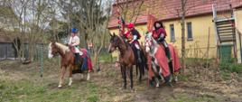 Na górze zdjęcia fragment nieba na tle którego znajdują się drzewa. W prawym górnym rogu czerwony dach oraz dom w kolorze żółtym. Na środku zdjęcia znajdują się trzy konie oraz sylwetki trzech osób siedzących na koniach. Sylwetki osób ubrane w różnokolorowe stroje historyczne z chorągwiami w dłoniach. Na dole zdjęcia trawa. Z lewej strony fragment budynku z czerwonym dachem.