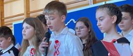 Uczeń klasy 7 odświętnie ubrany recytuje do mikrofonu na akademii, obok stoją inne odświętnie ubrane dzieci, trzymające w ręki biało-czerwone kartki