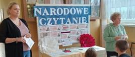 Pani wicedyrektor odczytuje fragment utworu Jana Kochanowskiego, obok stoi bibliotekarka szkolnej biblioteki, za paniami stół nakryty jasnym płótnem i gazetka informacyjna związana z imprezą z napisem "Narodowe Czytanie", na przodzie na małych ławeczkach siedzą słuchający uczniowie