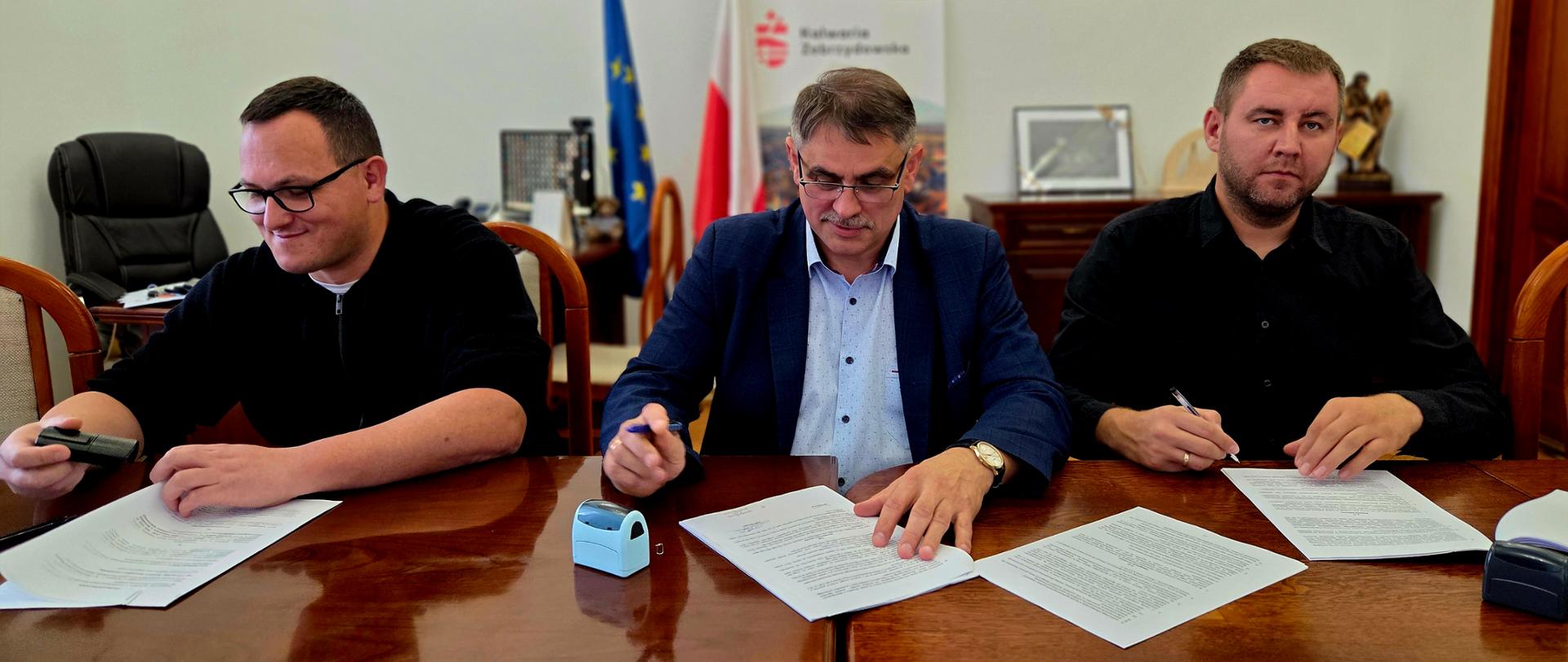 Trzech mężczyzn siedzących przy stole konferencyjnym w Urzędzie Miasta Kalwarii Zebrzydowskiej podczas podpisywania dokumentów związanych z realizacją inwestycji. W centralnej części fotografii przedstawiciel samorządu podpisuje umowę, a po jego obu stronach znajdują się pozostali uczestnicy spotkania. W tle widoczne są flagi Polski i Unii Europejskiej, godło państwowe oraz materiały promocyjne Gminy Kalwaria Zebrzydowska.