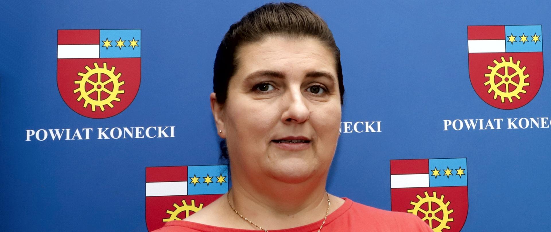 Fragment sylwetki kobiety przedstawiający głowę i ramiona. Kobieta ma ciemne oczy i ciemne włosy spięte gładko. W uszach ma drobne kolczyki z cyrkonią i złoty łańcuszek na szyi.