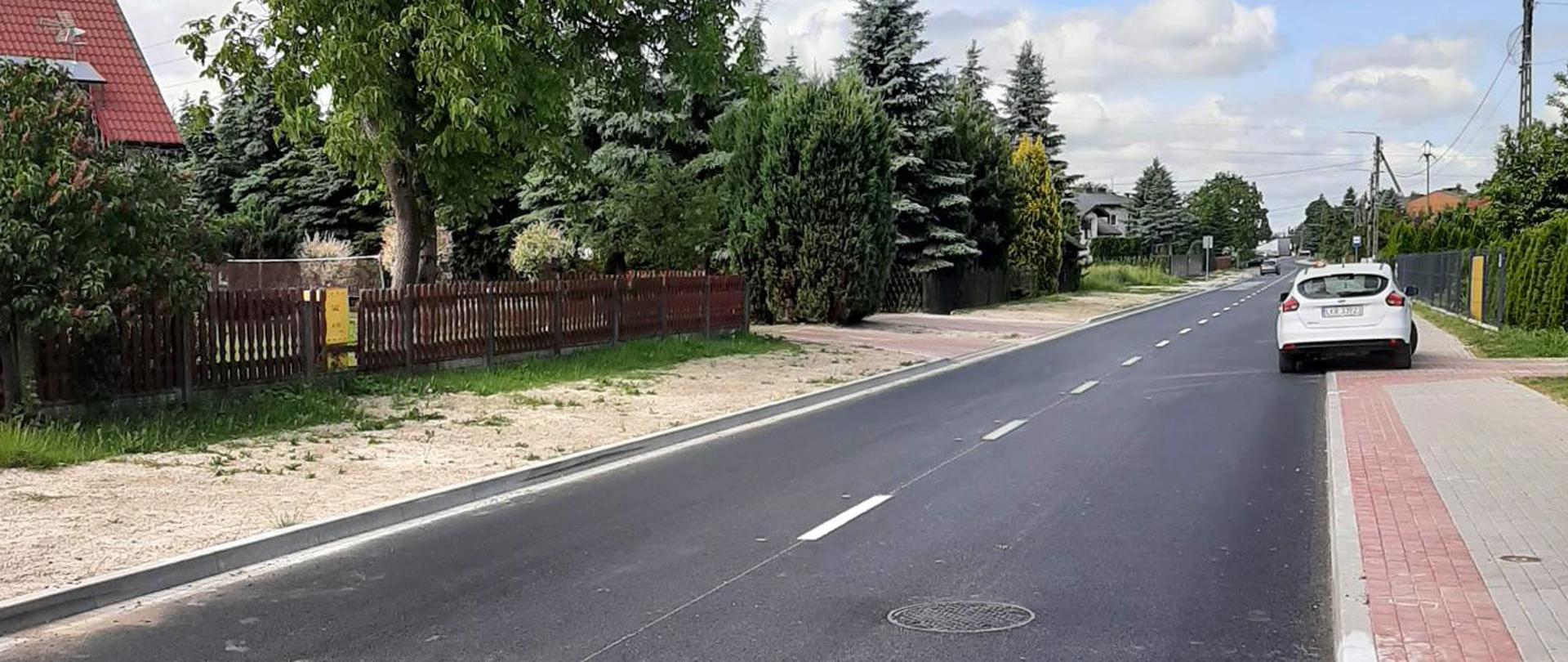 Zdjęcie przedstawia nawierzchnię nowo wyremontowanej drogi. Przy drodze znajduje się nowy chodnik z kostki brukowej przy którym stoi zaparkowane białe auto. W tle znajdują się drzewa i ogrody pobliskich zabudowań.