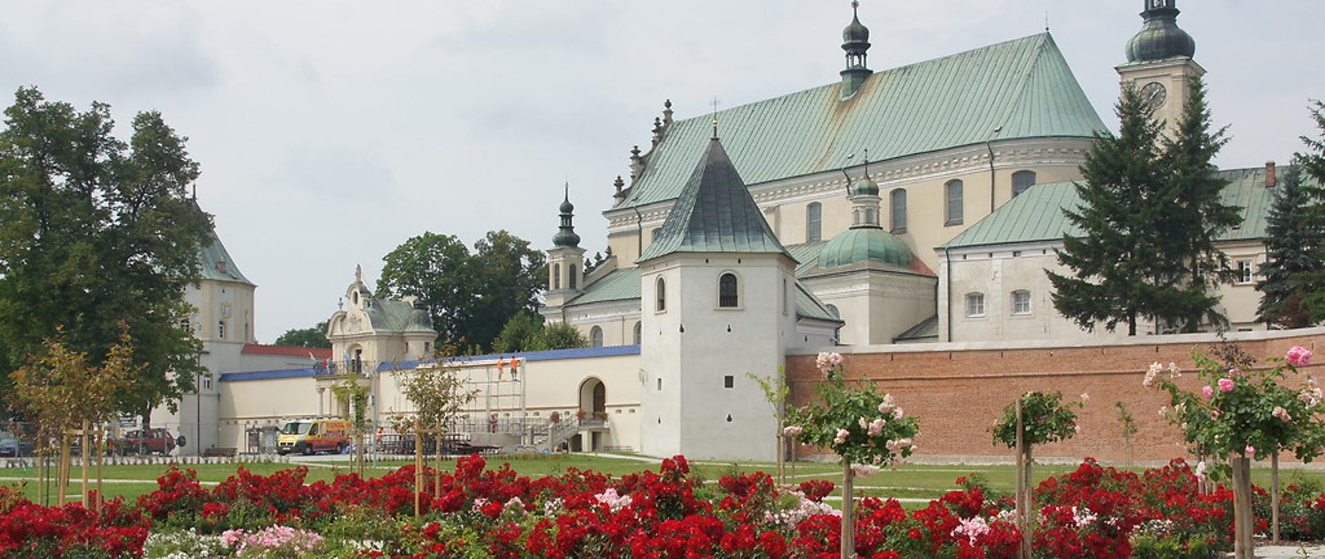 Klasztor otoczony grubym murem, przed klasztorem piękne rabaty kwiatów, głowienie czerwonych