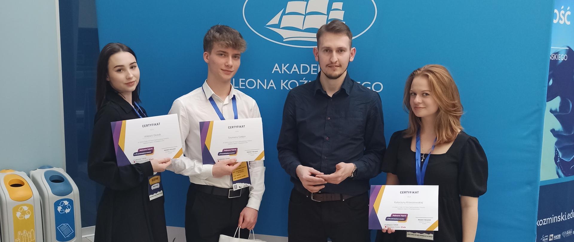 Uczniowie stoją przy ściance z nagrodami, które otrzymali w konkursie "Najlepsze Zajęcia z Przedsiębiorczości", który odbył się w Warszawie 