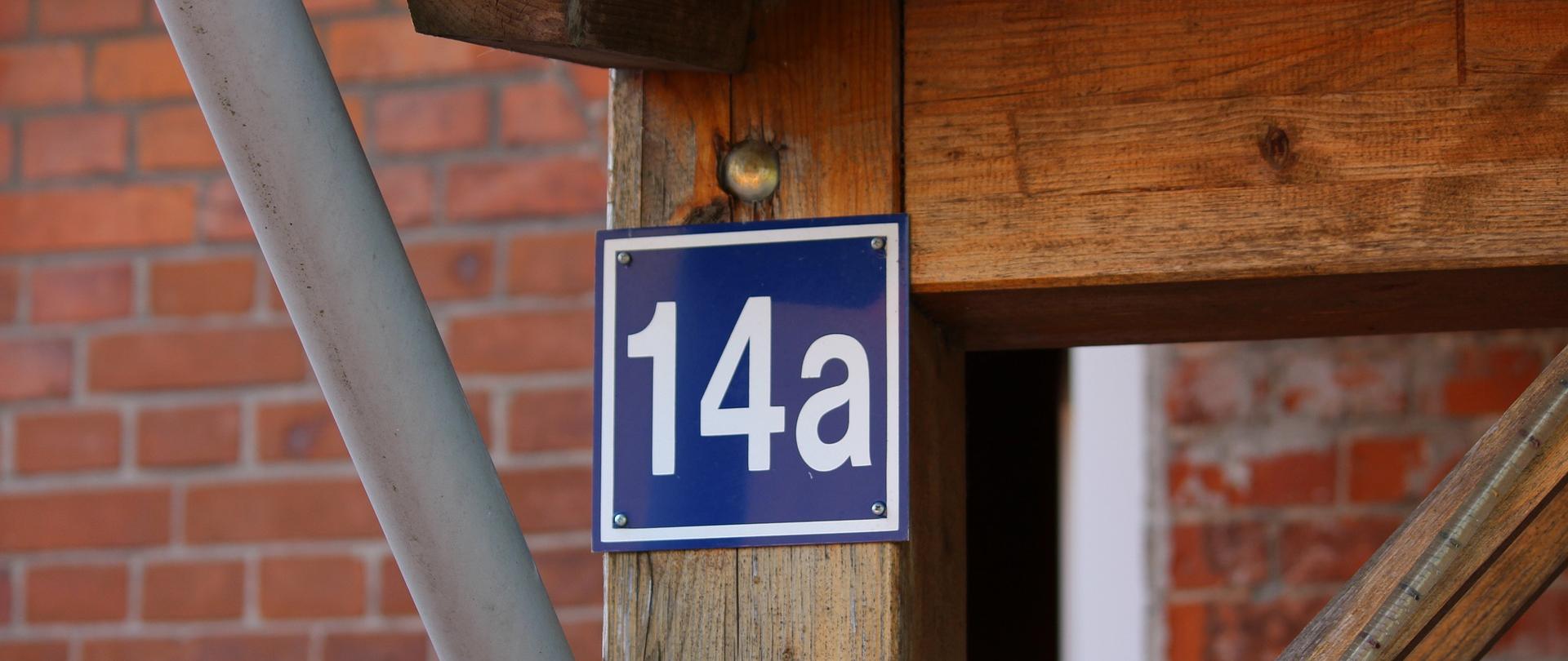 Tablica z numerem porządkowym budynku 14a przyczepiona do drewnianej konstrukcji przed ceglaną ścianą. Z konstrukcją łączy się także rynna.