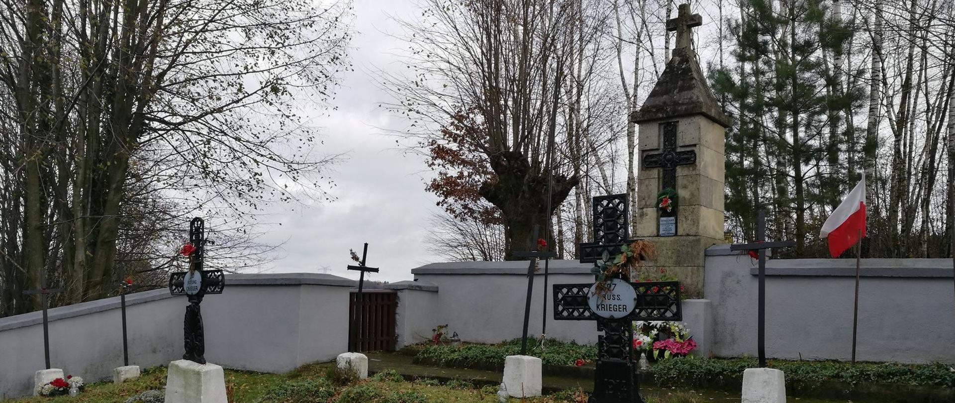 Zdjęcie przedstawia cmentarz wojskowy z kilkoma krzyżami i pomnikami. Cmentarz jest otoczony murem, a w tle widać drzewa i zachmurzone niebo. Na jednym z nagrobków można dostrzec napis "AUGUST KRIEGER", a także polską flagę. Na dole zdjęcia znajduje się podpis "Cmentarz wojenny nr 338 (Jasień) - 12.11.2017".