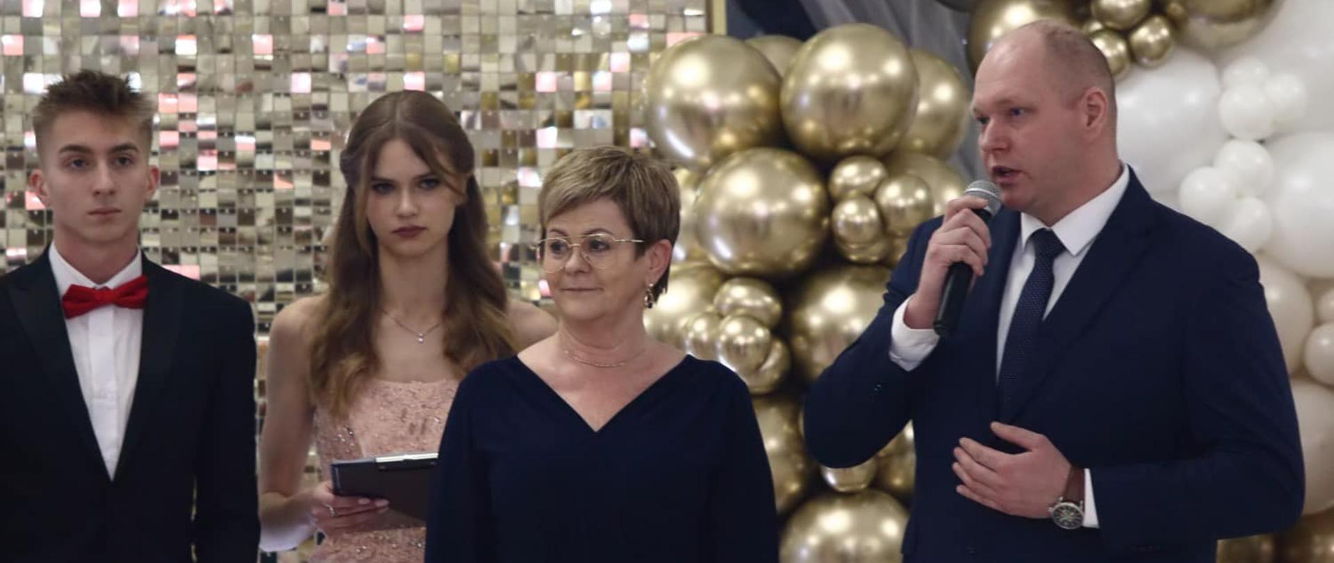Na zdjęciu widzimy oficjalny moment przemówienia podczas uroczystości, prawdopodobnie studniówki. Po prawej stronie znajduje się starosta gołdapski w eleganckim granatowym garniturze, trzymający mikrofon, co sugeruje, że jest w trakcie wygłaszania przemowy. Obok niego stoi wicestarosta w granatowej sukni, która uśmiecha się delikatnie.
Po lewej stronie widoczni są młodzi ludzie, pełniący prawdopodobnie funkcję prowadzących wydarzenie. Mężczyzna ma na sobie czarny garnitur z czerwonym krawatem w formie muszki, trzyma w rękach notatki. Obok niego stoi młoda kobieta w różowej, eleganckiej sukni, także z notatkami.
Tło zdobią efektowne dekoracje: złote balony oraz błyszcząca ścianka, które podkreślają odświętny i uroczysty charakter wydarzenia. Atmosfera jest pełna klasy i elegancji, typowa dla ważnych szkolnych uroczystości.