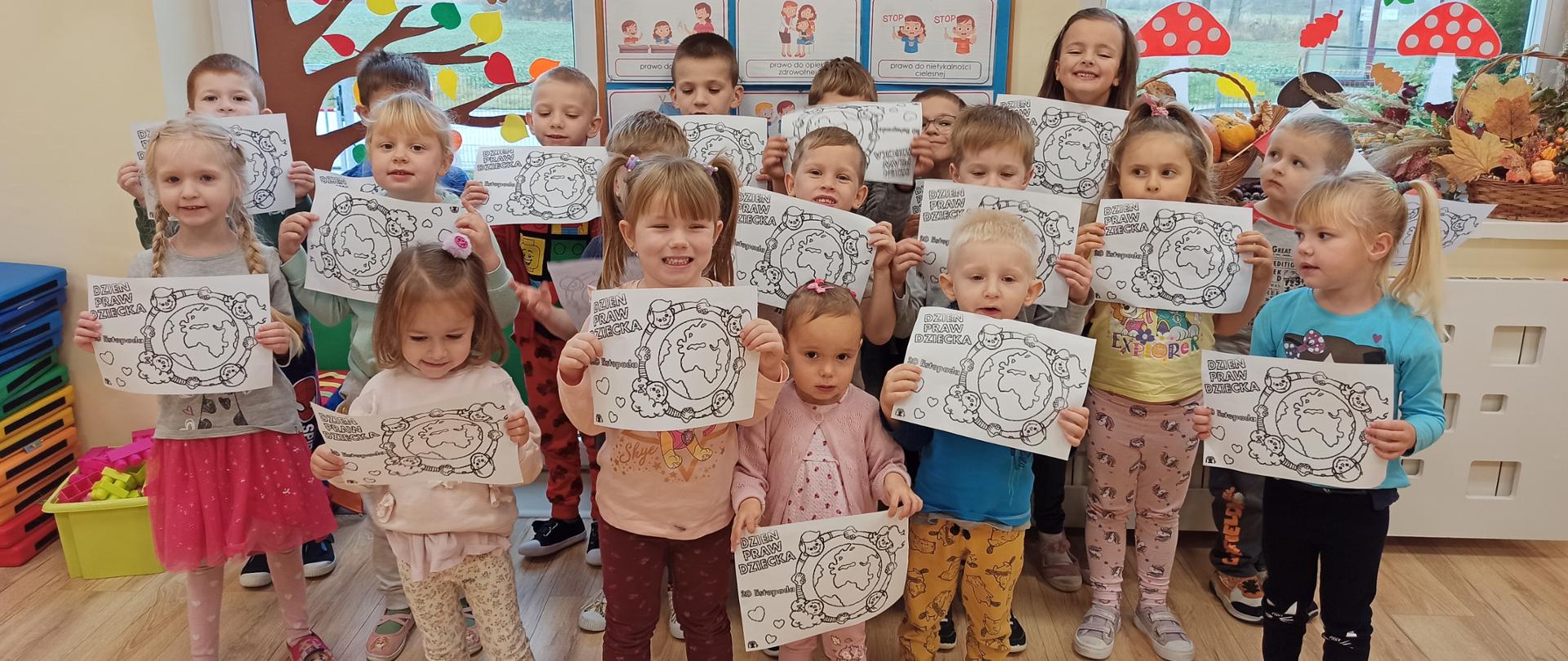Przedszkolaki stoją pod tablicą z prawami dziecka i pozują do zdjęcia z otrzymanymi kolorowankami.