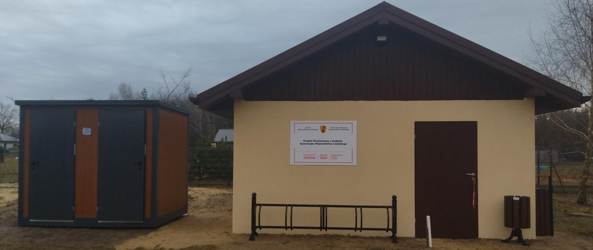 Budynek z dwuspadowym dachem oraz przylegającym do niego mniejszym pomieszczeniem gospodarczym. Fasada budynku jest beżowa, z brązowymi drzwiami, nad którymi znajduje się tablica informacyjna w języku polskim. Przed wejściem ustawiony jest stojak na rowery oraz kosz na śmieci. Obok stoi ciemnoszara szopa z brązowymi akcentami i dwoma drzwiami. Teren jest piaszczysty, a niebo zachmurzone.