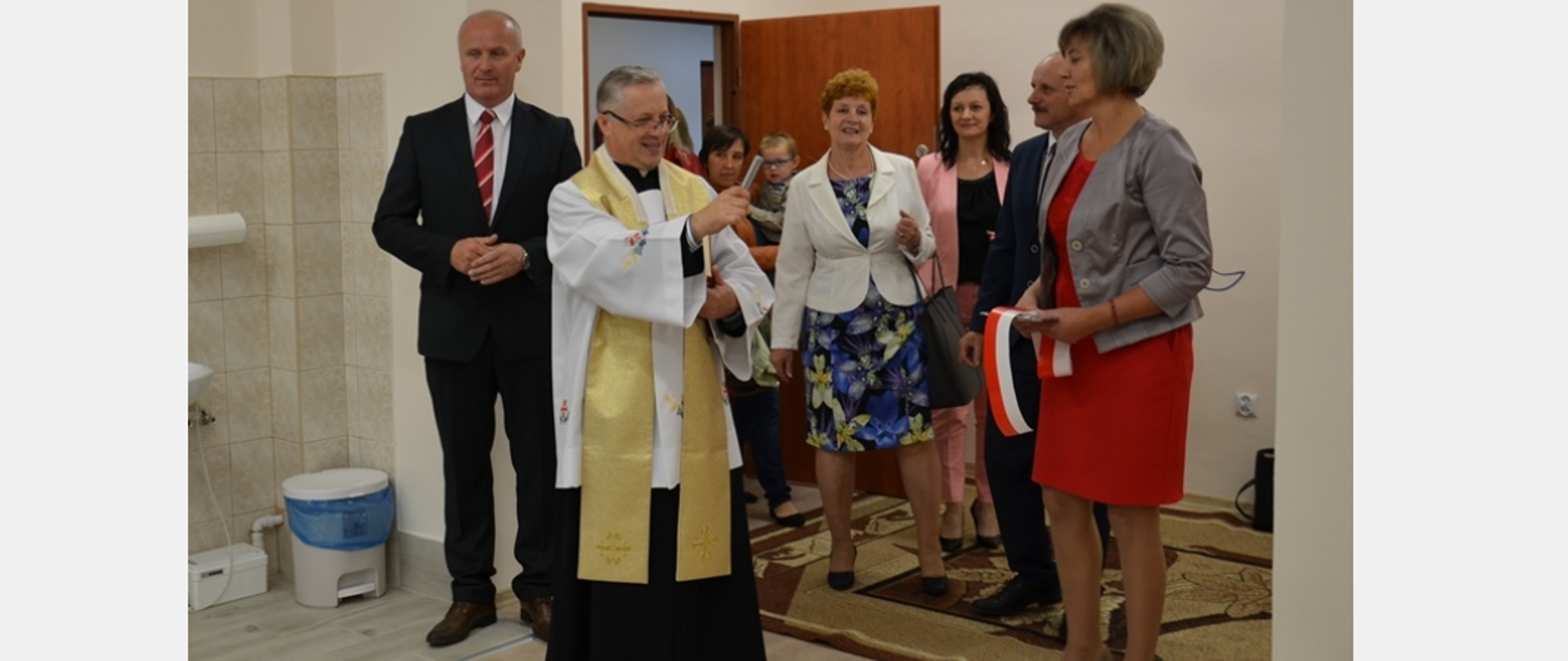 Na zdjęciu widzimy grupę ludzi, zdecydowanie wyróżnia się ksiądz który kropi pomieszczenie wodą święconą, na zdjęciu możemy zobaczyć również Pana Wójta oraz Panią Sekretarz 