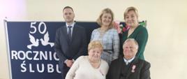 Państwo Maria i Stanisław na tle granatowej tablicy z napisem wraz z Wójtem, Z-cą Wójta i Z-cą Kierownika USC