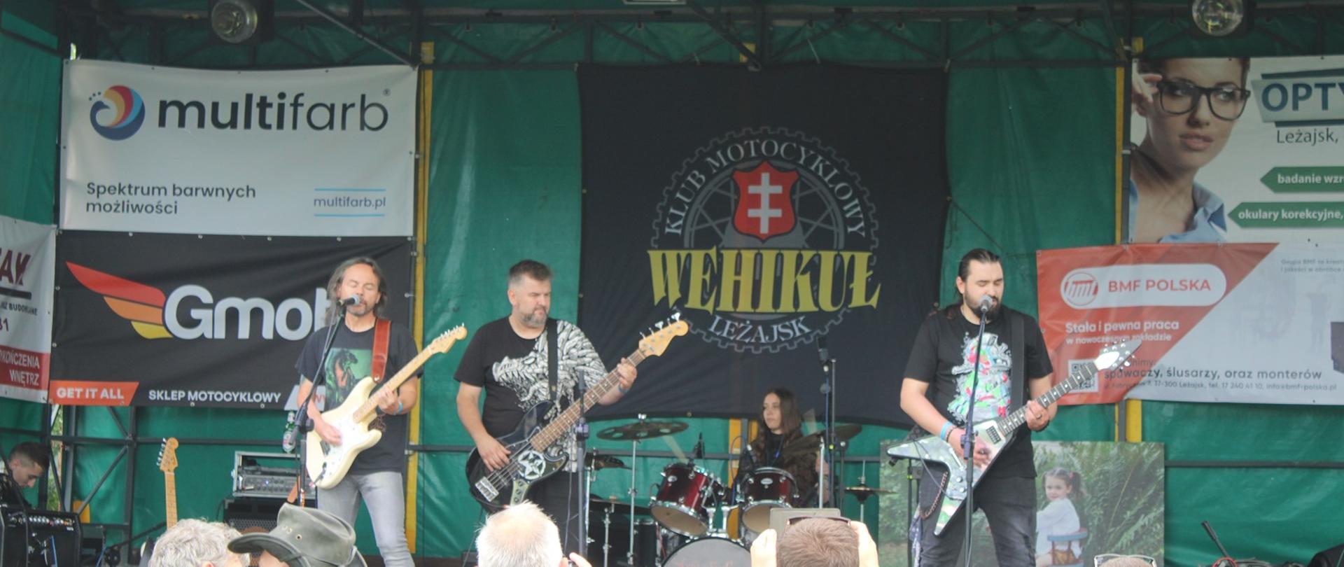 trzech mężczyzn z gitarami stoją na scenie, w tle kobieta gra na perkusji 