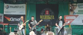 trzech mężczyzn z gitarami stoją na scenie, w tle kobieta gra na perkusji 