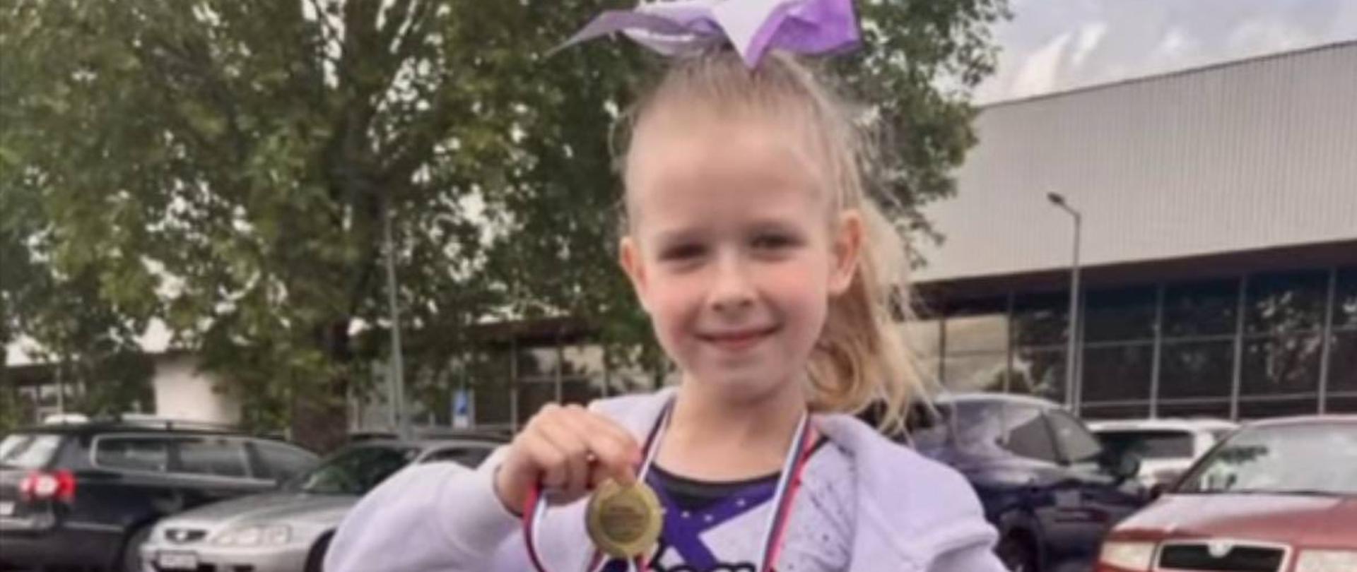 Ola ubrana w lawendowy stój cheerleaderki z fioletową kokardą na głowie trzyma w ręku złoty medal